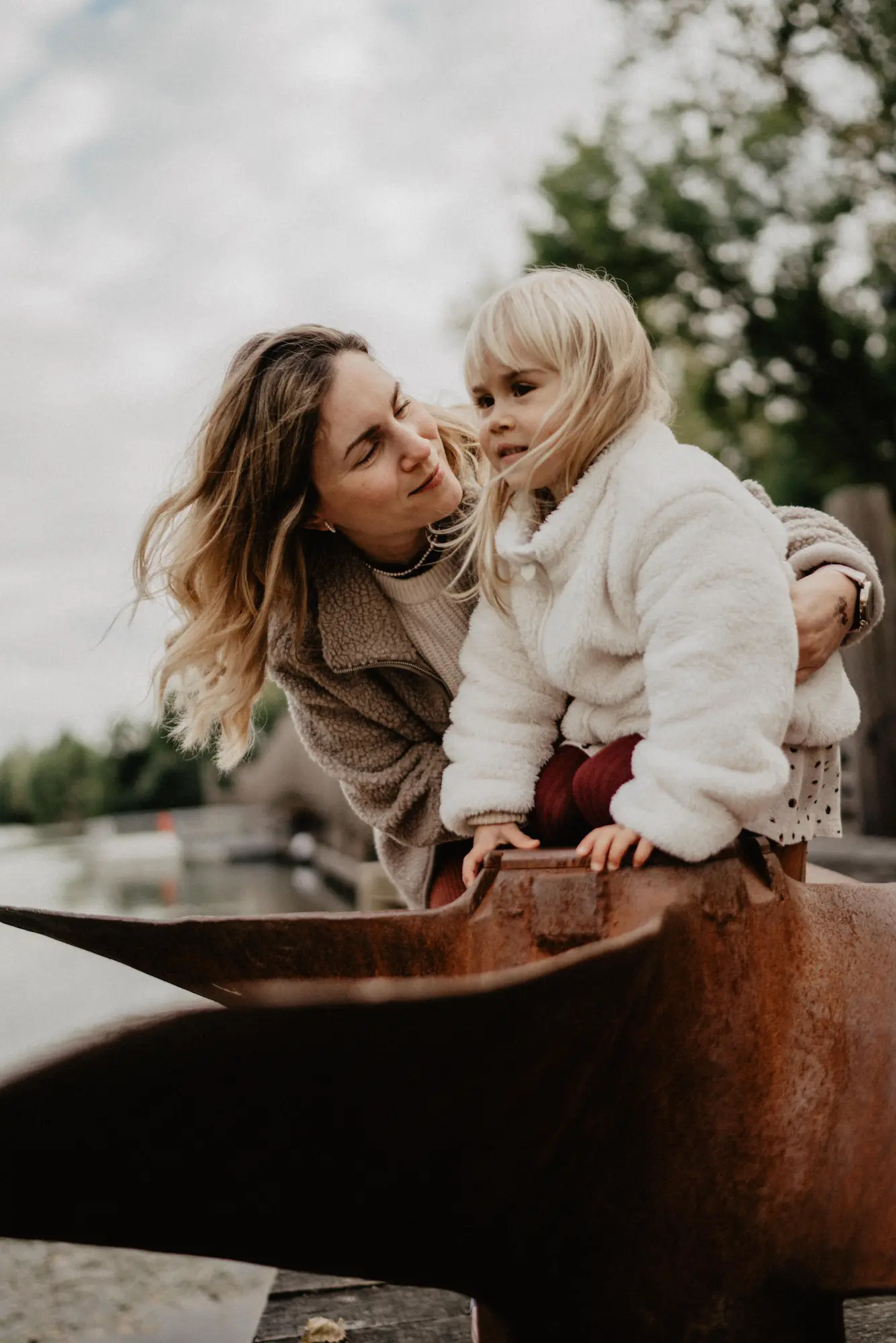 Mama sa nakláňa k malej blond dcérke pri hrdzavej soche pri rieke