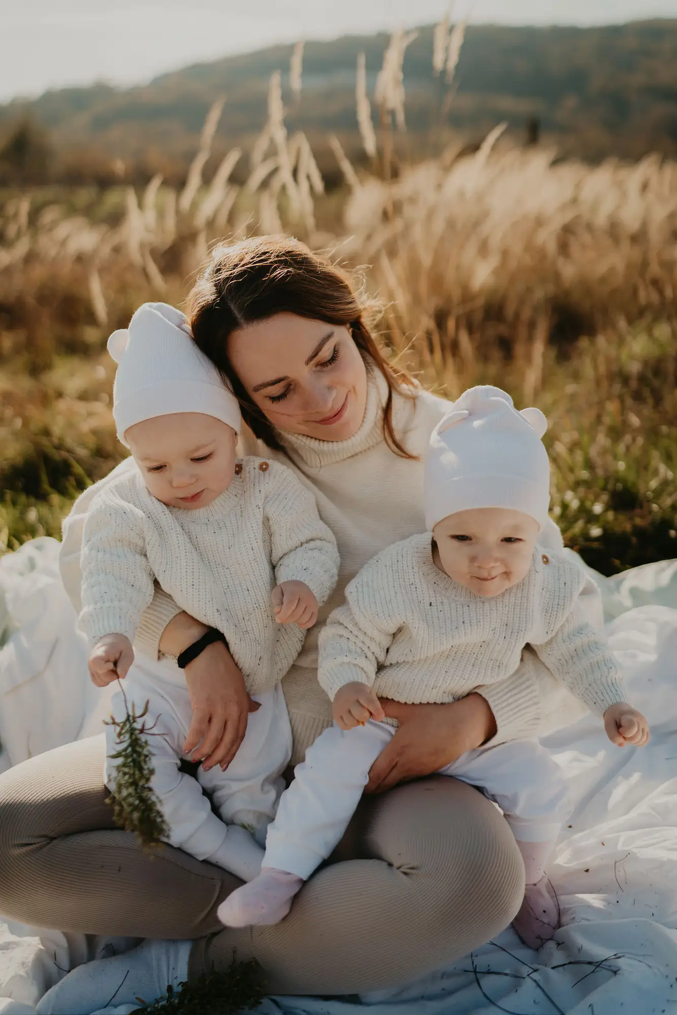 Mama s dvomi dvojičkami v bielych čiapočkách v poli s trávou v zlatom svetle