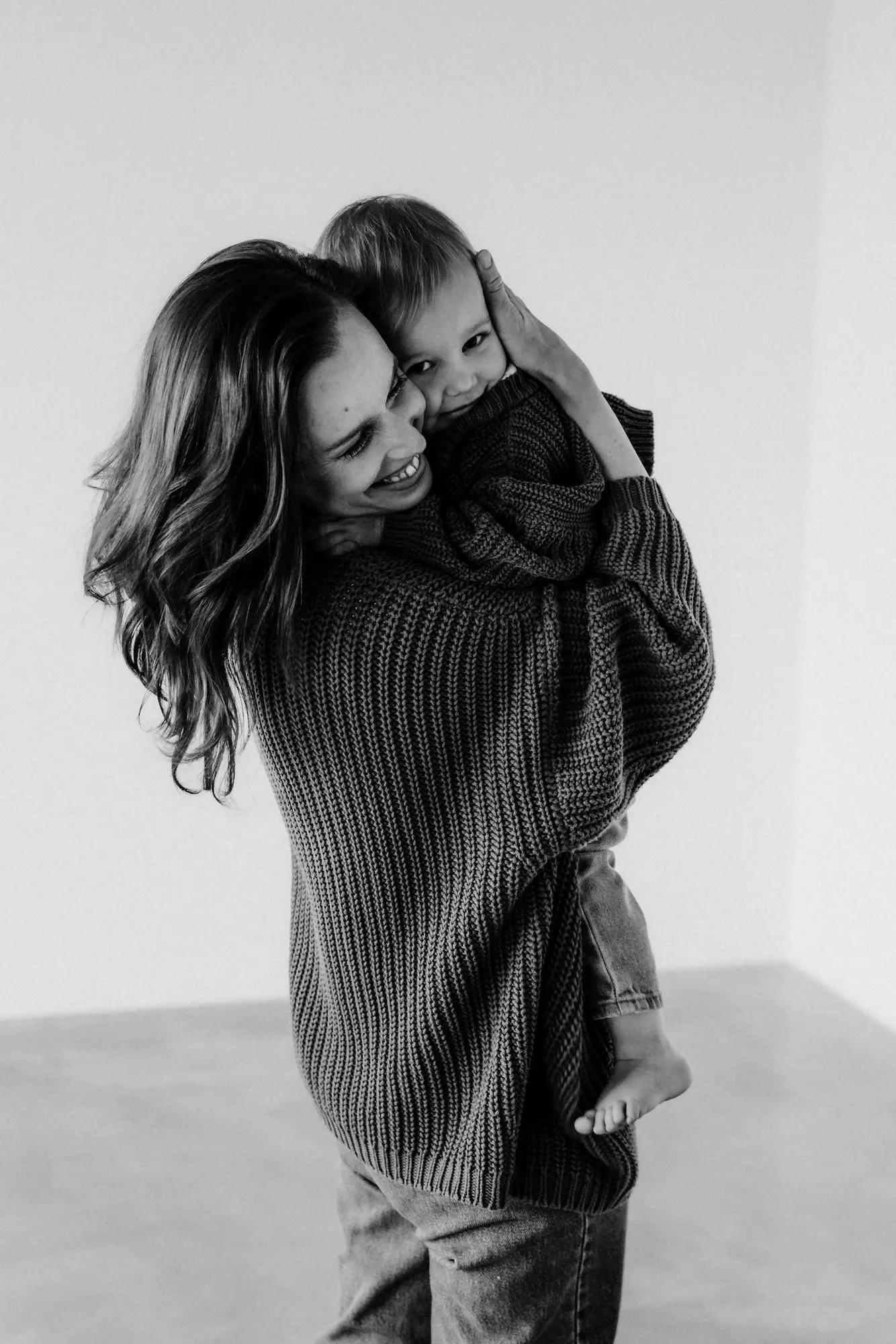 Mama nesie malé dieťa v pletenom svetri v štúdiu, čiernobiela fotografia
